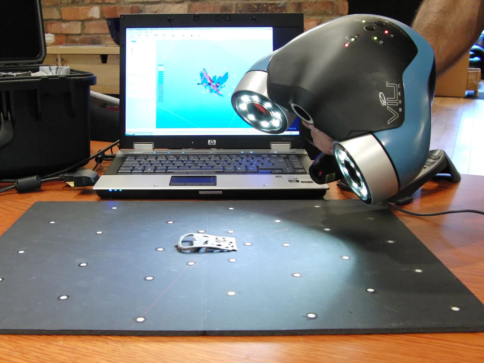 A handheld 3D scanner aimed at a belt buckle beside a laptop showing its digital model