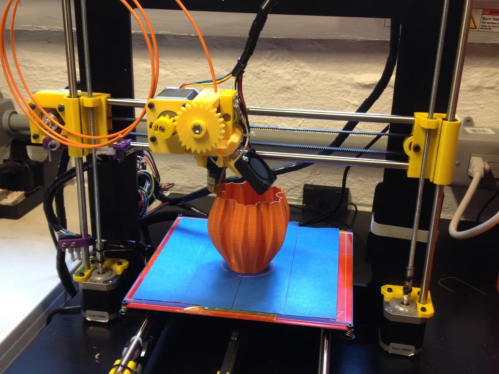 A filament 3D printer building an orange vase on a blue print bed
